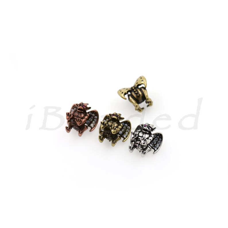 Peut inclure: Quatre petites figurines de gargouilles ail&eacute;es, chacune avec une finition diff&eacute;rente&nbsp;: cuivre, or, argent et bronze. Chaque gargouille a un petit bijou rose sur la poitrine.