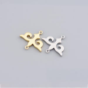 Peut inclure: Deux breloques décoratives en métal. L'une est dorée, l'autre argentée. Chaque breloque présente un motif symétrique stylisé avec des éléments incurvés et de petites boucles pour l'attache. Les breloques sont sur fond gris.
