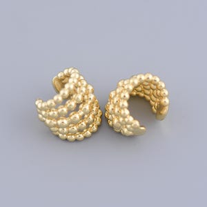 Może przedstawiać: Para złotych kolczyków w kształcie mankietów wykonanych z małych, okrągłych koralików. Mankietów są zaprojektowane tak, aby owijać się wokół ucha i wykonane są z wielu warstw koralików.