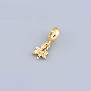 Puede incluir: Un charm dorado con un diseño de flor con cristales transparentes, amarillos y rosados. El charm tiene un bucle para sujetarlo a una pulsera o collar.