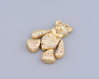 Puede incluir: Un dije de oso de peluche de tono dorado con diamantes de imitación transparentes. El oso tiene una pajarita y ojos negros.