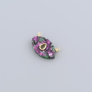 May include: An oval gemstone pendant with gold-coloured bails. The pendant is a mix of pink, green, and black. A smaller, teardrop-shaped gold-coloured accent is set in the centre. The pendant has two gold-coloured loops for attaching to a chain or other jewellery.