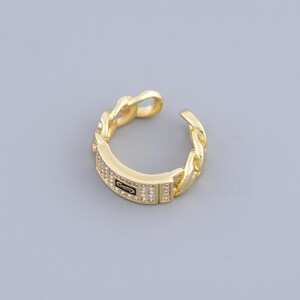 May include: Gold chain ring with a rectangular band encrusted with clear cubic zirconia stones. The band has a small black logo in the center.