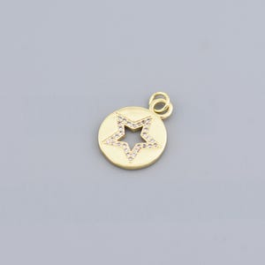 Puede incluir: Colgante dorado con un recorte en forma de estrella en el centro, bordeado con pequeñas piedras preciosas transparentes. El colgante circular tiene un lazo en la parte superior para sujetarlo a una cadena. Fondo gris liso.