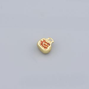 Puede incluir: Un dije de granada de color dorado con un grupo de piedras de color rojo anaranjado. El dije tiene un acabado liso y pulido y un pequeño lazo en la parte superior para sujetarlo a un collar o pulsera. El dije está sobre un fondo gris.