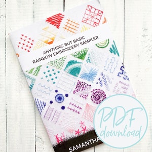 Può includere: Un libretto bianco con il titolo "Anything But Basic Rainbow Embroidery Sampler" e un ricamo colorato sulla copertina. Il libretto è su una superficie di legno bianco. Il testo "PDF download" è in un cerchio nell'angolo in basso a destra dell'immagine.