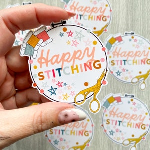 May include: A white round sticker with the text "Happy Stitching" in colourful letters. The sticker features a pair of gold scissors, a bird, and a variety of colourful stars.