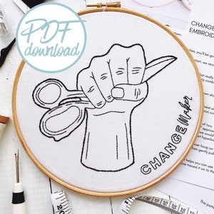 May include: A black and white embroidery pattern of a hand holding scissors with the text "CHANGE Maker" around the hand. The pattern is on a white fabric in a wooden embroidery hoop.