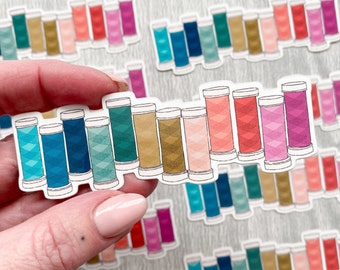 Könnte beinhalten: Eine Blatt mit bunten Fadenaufklebern in verschiedenen Farben wie Rosa, Rot, Blau, Grün und Gold. Die Aufkleber sind in Form von Garnrollen.