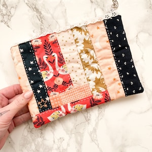 May include: A small quilted fabric pouch with a patchwork design featuring swans, stars, and geometric patterns in shades of pink, red, black, and white. The pouch has a white lace trim and a black zipper.