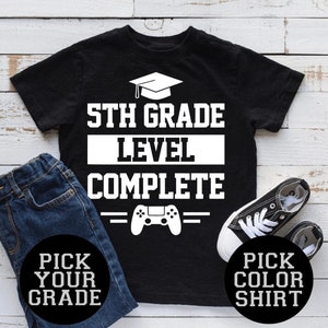 May include: Black t-shirt with white text that reads "5th Grade Level Complete" with a graduation cap and a game controller graphic. Includes a pair of blue jeans and black sneakers. The shirt also has the text "Pick Your Grade" and "Pick Color Shirt".