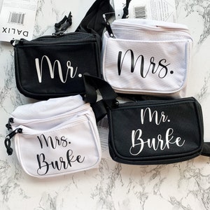 Op de afbeelding: Vier kleine, rechthoekige heuptasjes in zwart en wit. Elke tas heeft een hoofdvak met ritssluiting en een kleiner voorvak. De tassen zijn gelabeld met de woorden "Mr." en "Mrs." in wit script, met &eacute;&eacute;n tas die ook "Burke" leest.