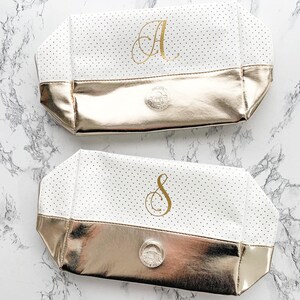 May include: Two white cosmetic bags with gold accents. The bags have a polka dot pattern and a gold metallic base. One bag has the letter "A" in gold, the other has the letter "S". Each bag has a silver emblem.