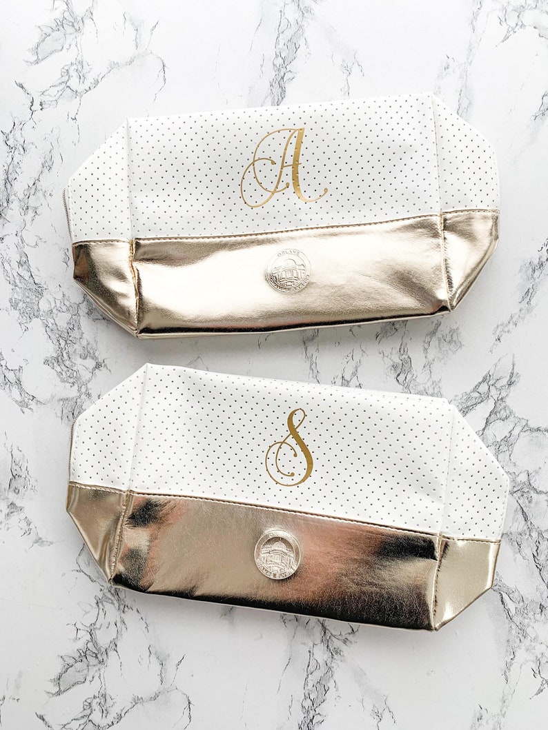 May include: Two white cosmetic bags with gold accents. The bags have a polka dot pattern and a gold metallic base. One bag has the letter "A" in gold, the other has the letter "S". Each bag has a silver emblem.