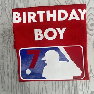 May include: Red t-shirt with the words "BIRTHDAY BOY" in white block letters. Below, a baseball-themed graphic features a silhouette of a batter and the number "7" in red, white, and blue.