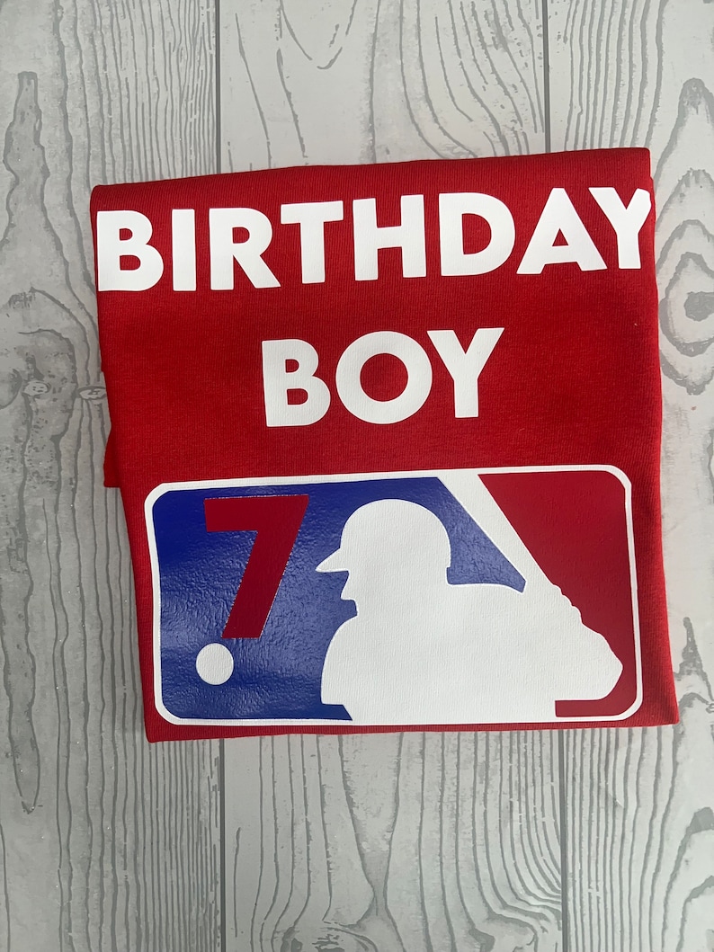 May include: Red t-shirt with the words "BIRTHDAY BOY" in white block letters. Below, a baseball-themed graphic features a silhouette of a batter and the number "7" in red, white, and blue.