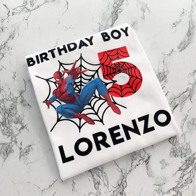 May include: White t-shirt with a red and blue Spiderman graphic, a black web design, and the text "Birthday Boy" and "Lorenzo" in black.