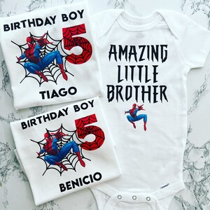 May include: Two white t-shirts with a Spiderman design. One shirt says "Birthday Boy" with the number 5 and the name "Tiago". The other shirt says "Birthday Boy" with the number 5 and the name "Benicio". A white onesie with a Spiderman design says "Amazing Little Brother".