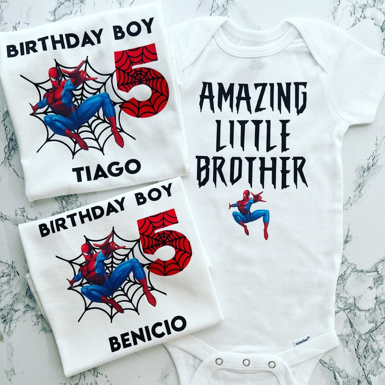 May include: Two white t-shirts with a Spiderman design. One shirt says "Birthday Boy" with the number 5 and the name "Tiago". The other shirt says "Birthday Boy" with the number 5 and the name "Benicio". A white onesie with a Spiderman design says "Amazing Little Brother".