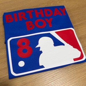 May include: Blue t-shirt with the words "BIRTHDAY BOY" in red. Features a baseball-themed graphic with the number "8" and a white silhouette of a batter. The shirt is folded on a wooden surface.