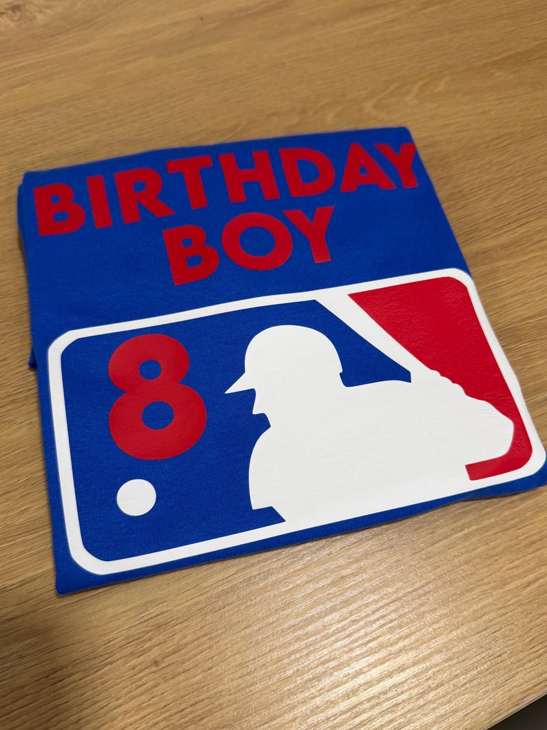 May include: Blue t-shirt with the words "BIRTHDAY BOY" in red. Features a baseball-themed graphic with the number "8" and a white silhouette of a batter. The shirt is folded on a wooden surface.