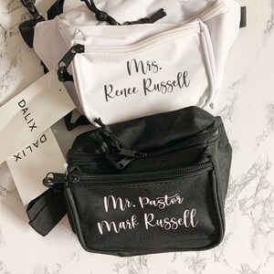 Op de afbeelding: Twee heuptasjes, &eacute;&eacute;n witte en &eacute;&eacute;n zwarte, worden getoond. Op het witte tasje staat "Mrs. Renee Russell" op de voorzak. Op het zwarte tasje staat "Mr. Pastor Mark Russell". Beide tasjes hebben zwarte ritsen en bandjes.