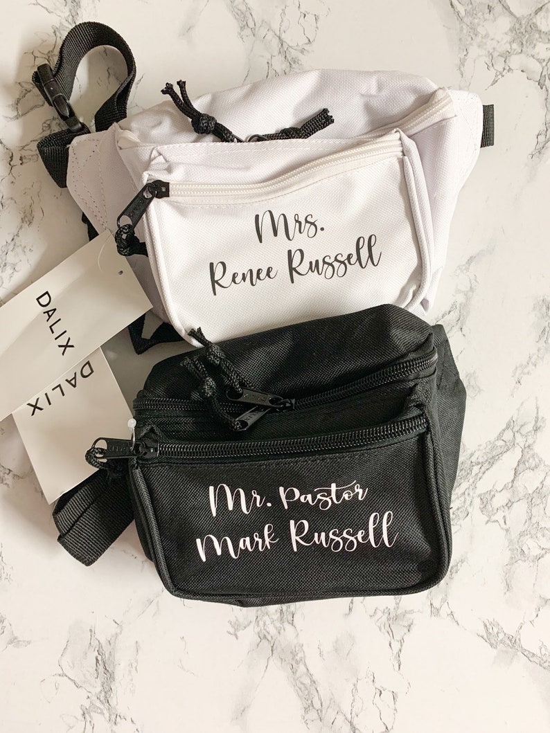 Op de afbeelding: Twee heuptasjes, &eacute;&eacute;n witte en &eacute;&eacute;n zwarte, worden getoond. Op het witte tasje staat "Mrs. Renee Russell" op de voorzak. Op het zwarte tasje staat "Mr. Pastor Mark Russell". Beide tasjes hebben zwarte ritsen en bandjes.