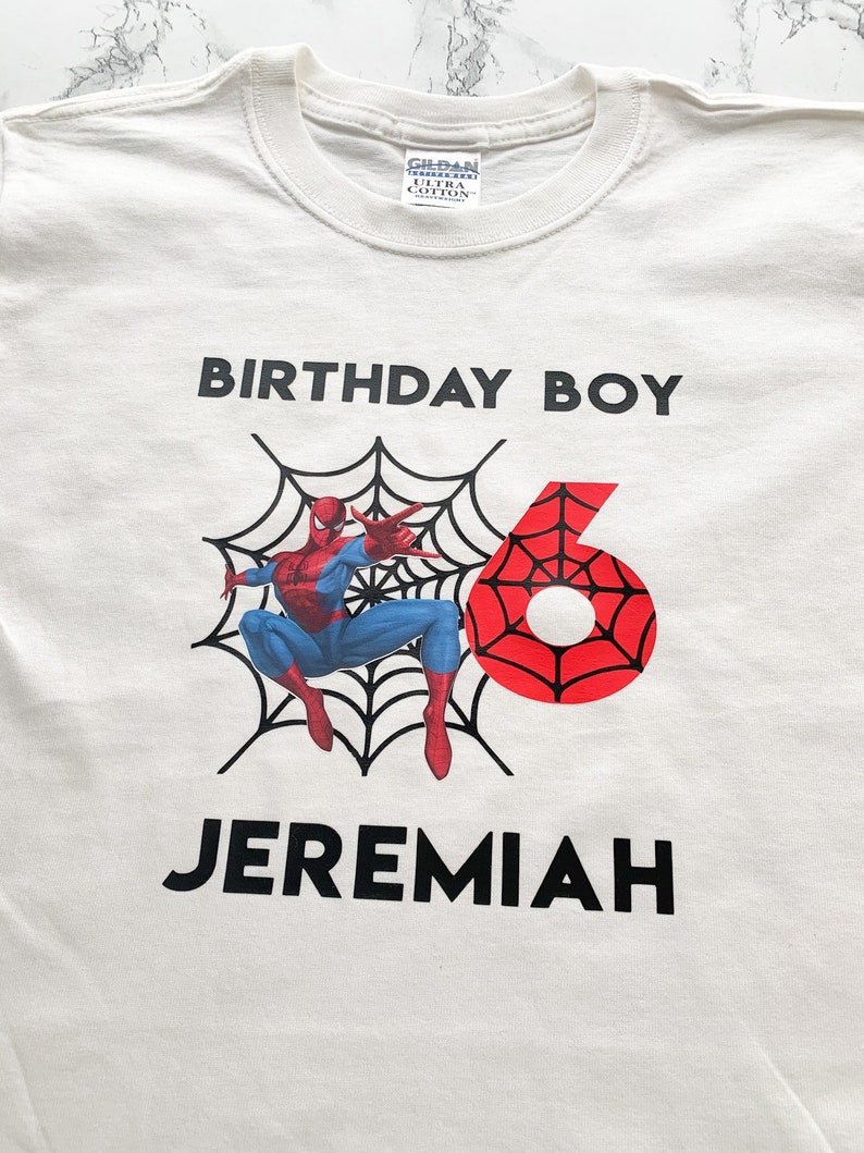 May include: White t-shirt with a red and blue Spiderman graphic, a black web, and the text "Birthday Boy" and "Jeremiah" in black.