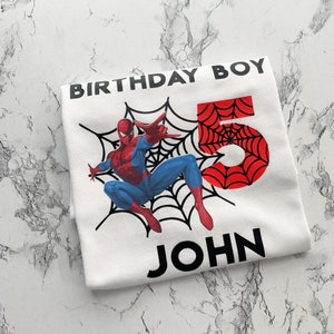 May include: White t-shirt with a red and blue Spiderman graphic, a black spiderweb, and the number 5. The text "Birthday Boy" and "John" are printed in black.