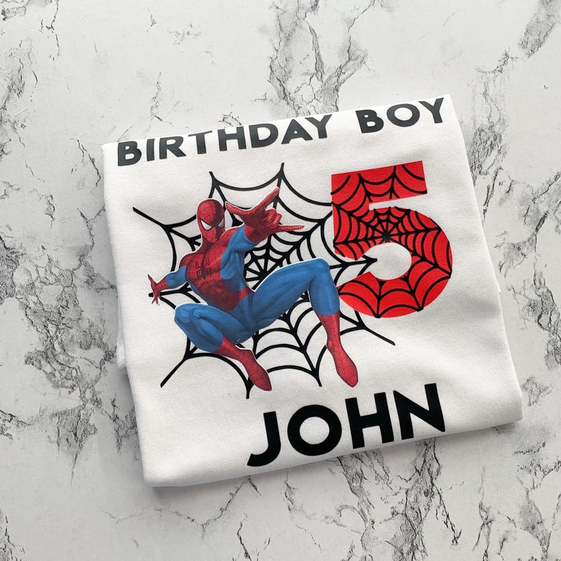 May include: White t-shirt with a red and blue Spiderman graphic, a black spiderweb, and the number 5. The text "Birthday Boy" and "John" are printed in black.