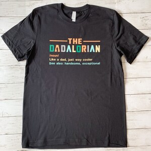 Op de afbeelding: Zwart T-shirt met een witte afbeelding met de tekst "THE DADALORIAN /noun/ Like a dad, just way cooler See also: handsome, exceptional"