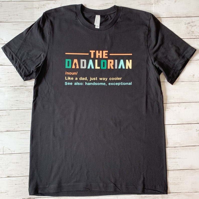 Op de afbeelding: Zwart T-shirt met een witte afbeelding met de tekst "THE DADALORIAN /noun/ Like a dad, just way cooler See also: handsome, exceptional"