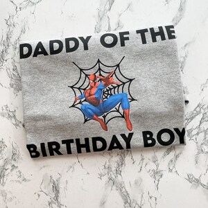 May include: Gray t-shirt with black text that reads "Daddy of the Birthday Boy". A cartoon image of Spiderman in a web is centered on the shirt.