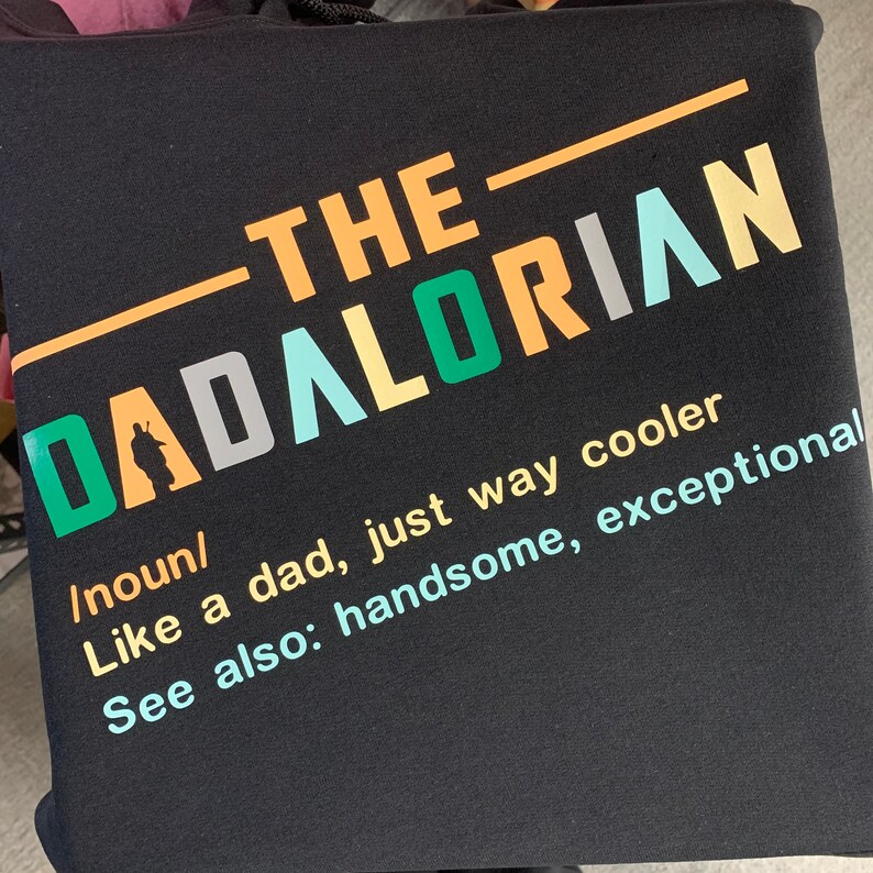 Op de afbeelding: Zwarte hoodie met een witte, groene en oranje afbeelding met de tekst "The Dadalorian" en de definitie als zelfstandig naamwoord, "Net als een vader, alleen veel cooler." Er staat ook "Zie ook: knap, uitzonderlijk."