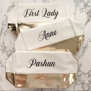 May include: Three white cosmetic bags with gold accents and black polka dots. The bags have the names "First Lady", "Anne", and "Pashun" printed on them in elegant script. The bottom of each bag is gold.