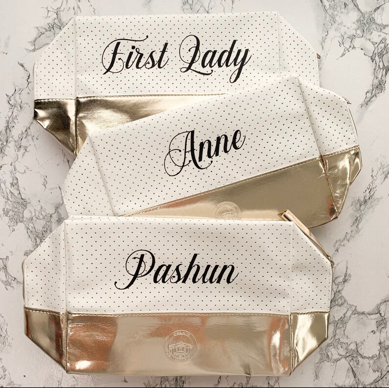 May include: Three white cosmetic bags with gold accents and black polka dots. The bags have the names "First Lady", "Anne", and "Pashun" printed on them in elegant script. The bottom of each bag is gold.