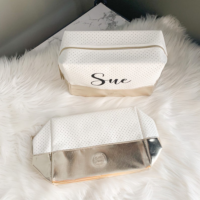 May include: Two white cosmetic bags with gold accents. The bags have a perforated design and a gold zipper. The larger bag has the name "Sue" printed on it. The smaller bag has a gold base with a circular emblem.