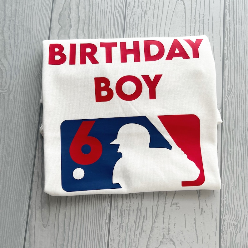 May include: White t-shirt with the words "BIRTHDAY BOY" in red block letters. Below, a baseball-themed graphic with a silhouette of a batter, the number "6", and red and blue accents. The shirt is folded.