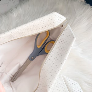 May include: A white, perforated case with a gold zipper, open to reveal a pair of scissors. The scissors have gray handles with yellow accents and long, silver blades. The case is on a white, fluffy surface.