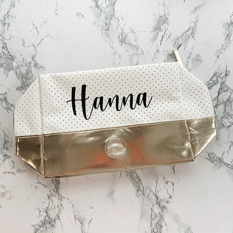 May include: A white cosmetic bag with black polka dots and a gold metallic bottom. The name "Hanna" is written in black script across the top. A small silver emblem is on the gold bottom.