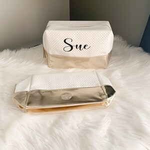 May include: Two white cosmetic bags with gold trim. The larger bag is rectangular with the name "Sue" printed on it. The bags have a perforated design and a gold base. The bags are on a white, fluffy surface.