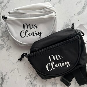 Op de afbeelding: Twee heuptasjes, &eacute;&eacute;n witte en &eacute;&eacute;n zwarte, met de namen "Mrs. Cleary" en "Mr. Cleary" in witte letters. Beide tassen hebben een hoofdvak met ritssluiting en verstelbare bandjes. De achtergrond is een marmerpatroon.