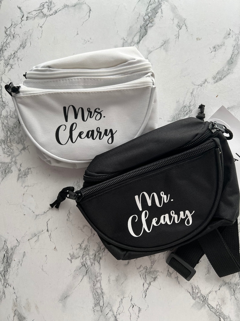 Op de afbeelding: Twee heuptasjes, &eacute;&eacute;n witte en &eacute;&eacute;n zwarte, met de namen "Mrs. Cleary" en "Mr. Cleary" in witte letters. Beide tassen hebben een hoofdvak met ritssluiting en verstelbare bandjes. De achtergrond is een marmerpatroon.