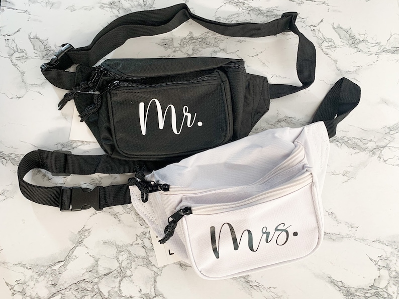 Op de afbeelding: Twee heuptassen, &eacute;&eacute;n zwarte en &eacute;&eacute;n witte, worden getoond. De zwarte tas heeft "Mr." in witte letters op de voorkant, terwijl de witte tas "Mrs." in dezelfde letters heeft. Beide hebben verstelbare zwarte bandjes en ritsen.