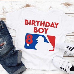 May include: White t-shirt with the text "BIRTHDAY BOY" in red. The shirt features a baseball-themed graphic with the number "8" in red. The image also includes blue jeans and white sneakers.