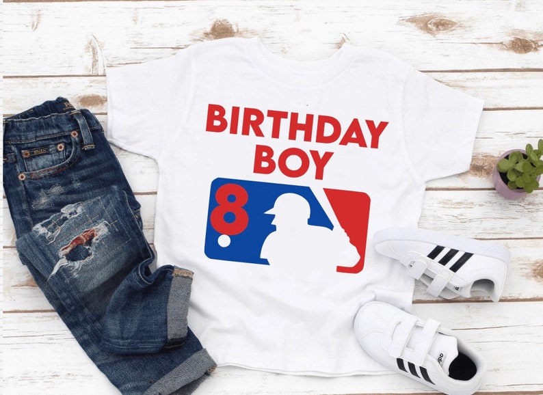 May include: White t-shirt with the text "BIRTHDAY BOY" in red. The shirt features a baseball-themed graphic with the number "8" in red. The image also includes blue jeans and white sneakers.