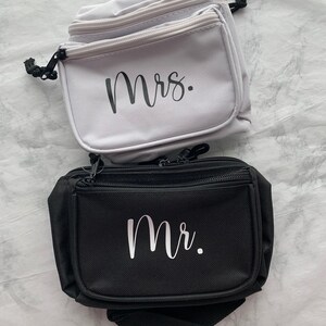 Op de afbeelding: Twee heuptasjes, &eacute;&eacute;n lichtgrijs en &eacute;&eacute;n zwart, worden getoond. Op het lichtgrijze tasje staat het woord "Mrs." in zwart schrift. Op het zwarte tasje staat het woord "Mr." in wit schrift. Beide hebben ritsen en zwarte trekkoorden.
