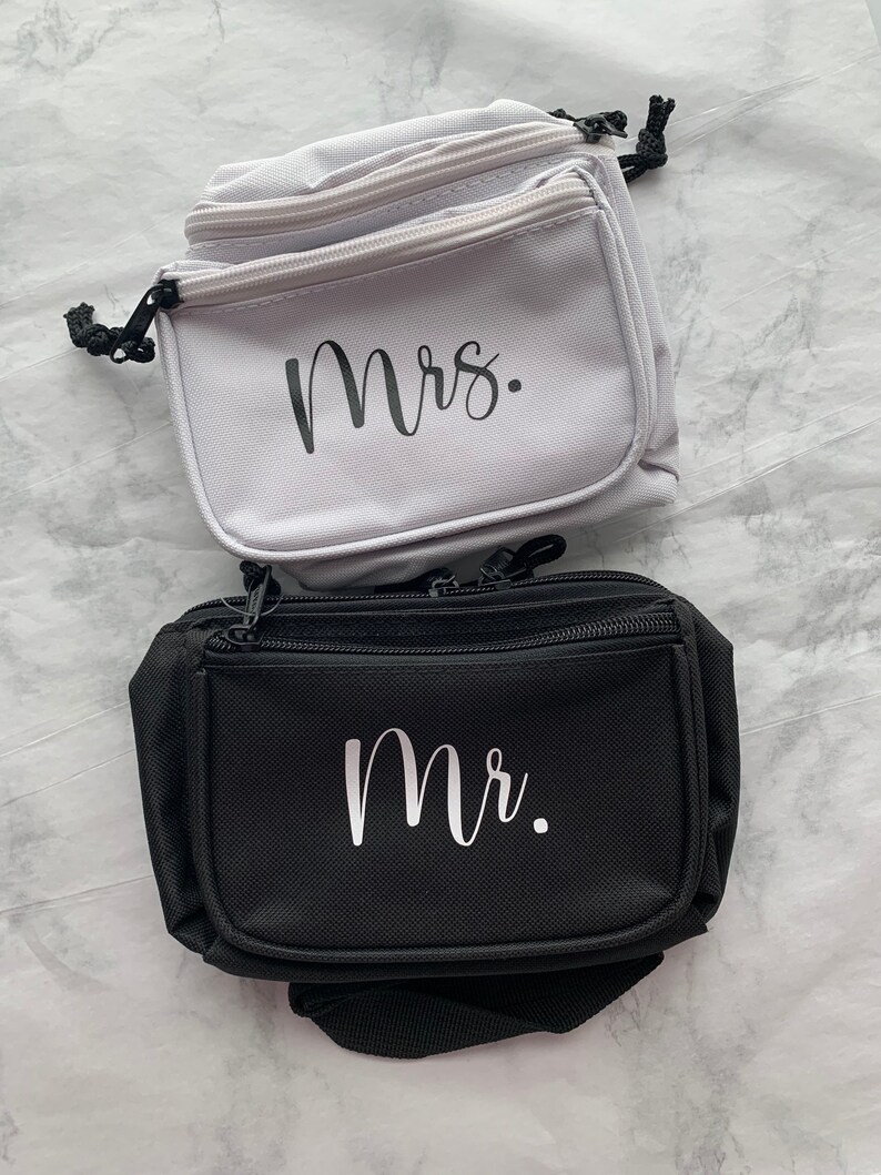 Op de afbeelding: Twee heuptasjes, &eacute;&eacute;n lichtgrijs en &eacute;&eacute;n zwart, worden getoond. Op het lichtgrijze tasje staat het woord "Mrs." in zwart schrift. Op het zwarte tasje staat het woord "Mr." in wit schrift. Beide hebben ritsen en zwarte trekkoorden.