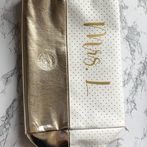 May include: A two-toned cosmetic bag, half gold and half white with small perforations. The gold side has a circular emblem. The white side has the gold text "Mrs. L" written vertically. The bag has a zipper closure.