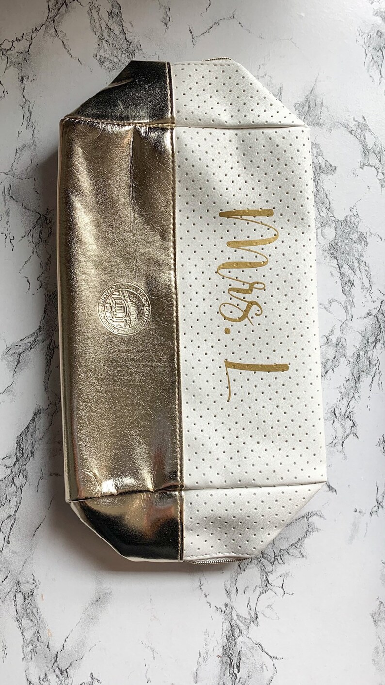 May include: A two-toned cosmetic bag, half gold and half white with small perforations. The gold side has a circular emblem. The white side has the gold text "Mrs. L" written vertically. The bag has a zipper closure.